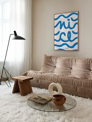 Soggiorno accogliente con divano beige, lampada da terra nera, tavolino in vetro e poster blu