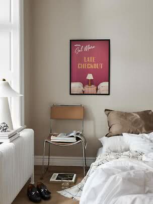 Soveværelse med uredt seng, stol med magasiner og plakat med teksten "Late Checkout" på væggen
