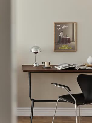Skrivebord i mørkt træ med kromlampe, kop, åben bog og sort stol ved væg med indrammet plakat