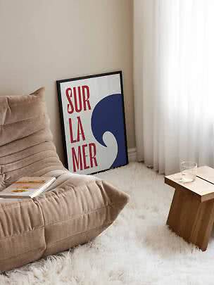 Rincón acogedor con sillón beige, mesa auxiliar de madera y póster Sur La Mer enmarcado