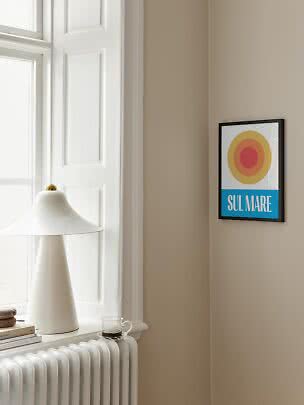 Framed SUL MARE poster with colored circles on a beige wall beside a window and white lamp