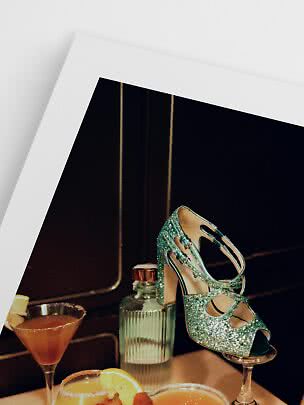 Glittery turquoise high-heeled sandal on a cocktail glass beside drinks and a bottle