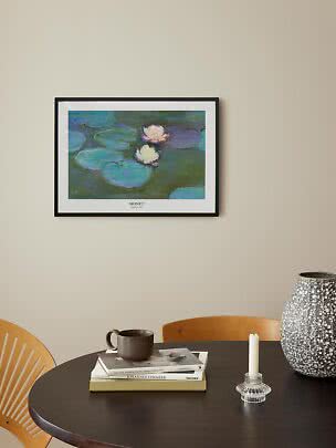 Lámina enmarcada de Monet con nenúfares sobre mesa oscura con libros, taza, candelero y jarrón grande.