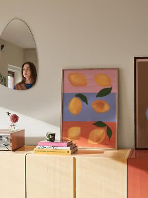 Farverig plakat med citroner i træramme, lænet på lys skænk i solrigt rum