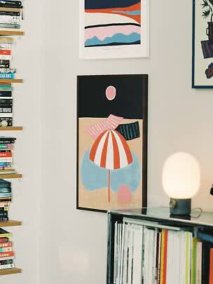 Framed abstract poster on a wall above a bookshelf with books and a small lamp