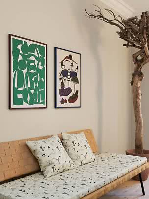 Woven daybed with patterned mattress and cushions beneath two framed posters on a light wall