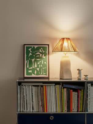 Tischlampe mit plissiertem Schirm auf Bücherregal neben grün gerahmtem Bild und kleinen Vasen