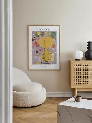 Framed Hilma af Klint poster on a beige wall in a living room with a white armchair and wood cabinet