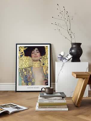 Inramad Gustav Klimt-affisch mot väggen med böcker, mugg och svart vas på vit piedestal