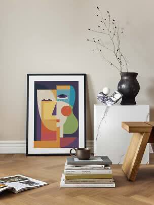 Framed abstract face poster leaning by a wall beside a marble pedestal with vase, books and mug