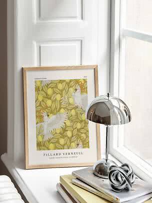 Framed botanical bird print on a white windowsill beside a chrome table lamp and stacked books
