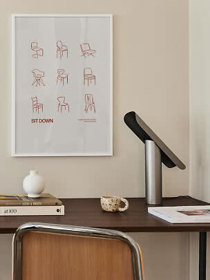 Minimalistisk skrivebord med sort bordlampe, bøger, vase og indrammet stolplakat på væggen