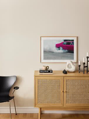Helles Sideboard mit Geflechtüren, schwarzer Stuhl und gerahmtes Foto eines pinken Autos an beiger Wand