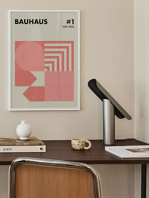 Skrivebord med sort bordlampe og indrammet Bauhaus-plakat i rosa toner på væggen