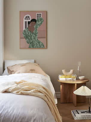 Schlafzimmer mit weißem Bett, Holztisch und gerahmtem Kunstdruck einer Frau an beigefarbener Wand