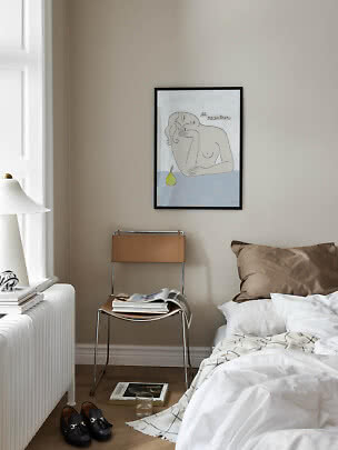 Schlafzimmer mit Bett, Stuhl, Büchern und gerahmtem Kunstdruck an beigefarbener Wand