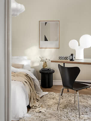 Lyst soveværelse med hvid seng, sort stol, skrivebord og stor hvid bordlampe