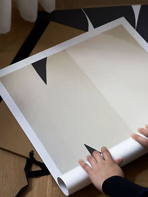 Hænder ruller en abstrakt plakat i beige, sort og brun på et trægulv