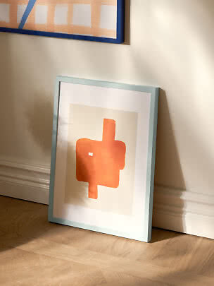 Indrammet abstrakt print med orange motiv, lænet mod en beige væg i sollys