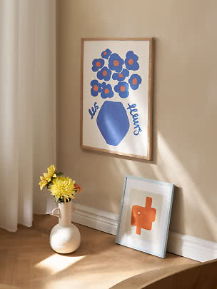 Ingelijste poster met blauwe bloemen in blauwe vaas aan beige muur, naast vaas met gele bloemen.