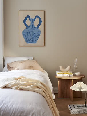 Gemütliches Schlafzimmer mit weißem Bett, Holznachttisch und blauem Vasenbild an beiger Wand
