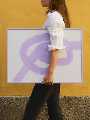 Woman in white shirt carrying a large framed abstract print with lilac brushstrokes against a yellow wall