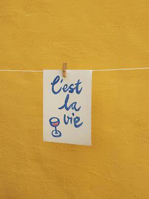 White card with blue "C'est la vie" text and wine glass, clipped to a line against a yellow wall