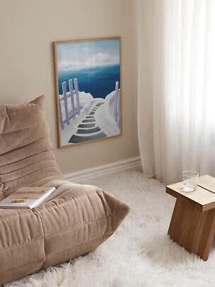 Cuadro enmarcado de un sendero blanco junto al mar azul, en pared beige junto a un sillón