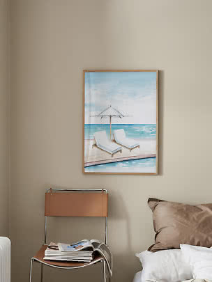 Framed beach print with two lounge chairs and umbrella by the sea on a beige wall