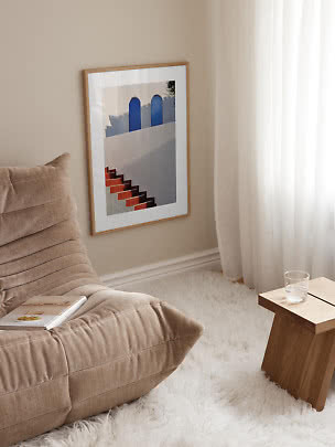 Framed art print with blue arches and red stairs on a beige wall beside a lounge chair