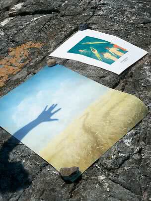 To kunstplakater ligger på klipper udendørs, holdt nede med sten.