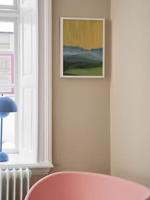Framed landscape painting with blue mountains and green fields on a beige wall