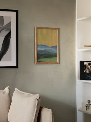 Small framed landscape painting with blue mountains and yellow sky on a green wall