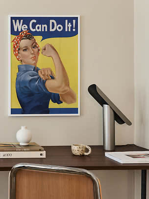 Framed 'We Can Do It!' poster above desk with lamp, books, vase and mug