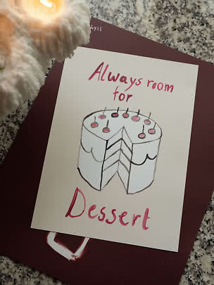 Carte avec dessin de gâteau à étages et le texte « Always room for dessert » près d’une bougie allumée.