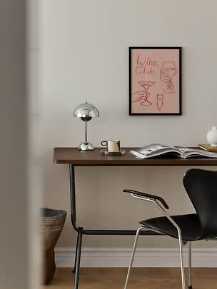 Scrivania con lampada cromata, tazza, libro aperto e sedia nera sotto un poster rosa Wine Club