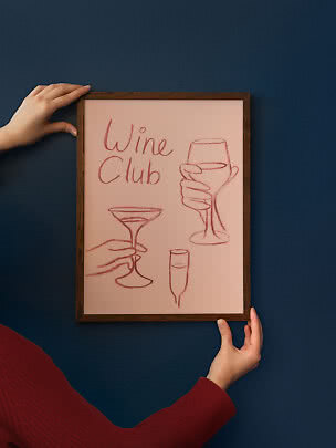 Poster rosa incorniciato Wine Club con schizzi di calici, tenuto contro una parete blu scuro
