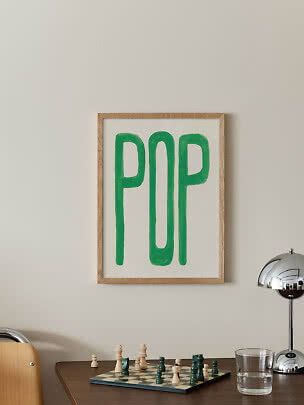 Framed poster with green 'POP' lettering on a light wall above a table with a chessboard.