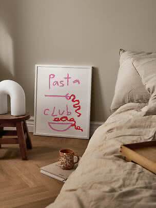 Indrammet Pasta Club-plakat lænet mod væg ved seng med beige sengetøj og lille træskammel