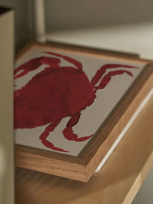 Framed print of a red crab on a white background, resting on a wooden shelf