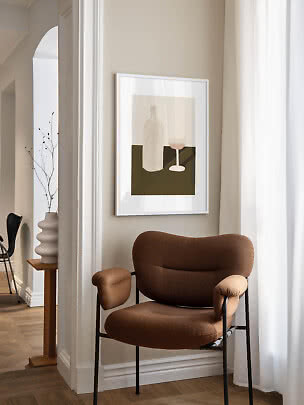 Brun polstret stol ved beige væg med indrammet stillebenplakat og hvid gardin