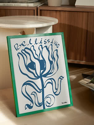 Ingelijste poster met blauwe bloem en de tekst "Bellissimo", leunend tegen een tafel in houtinterieur