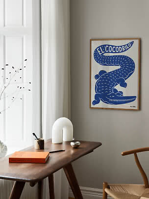 Indrammet plakat med blå krokodille på beige væg over et træbord ved et vindue