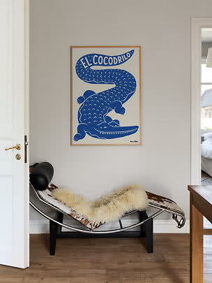 Blå plakat med krokodille og teksten "El Cocodrilo" i lys træramme over loungestol.