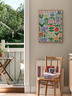 Gerahmtes FLORA-Blumenposter an der Wand über einem Holzstuhl in hellem Raum