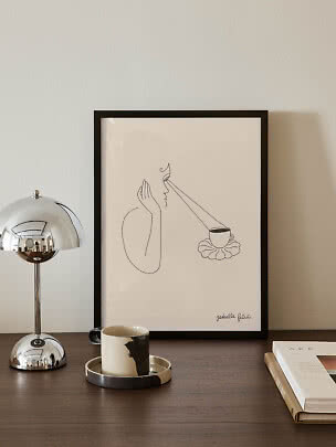 Gerahmte minimalistische Linienzeichnung auf Tisch mit Chromlampe, Tasse und Buch