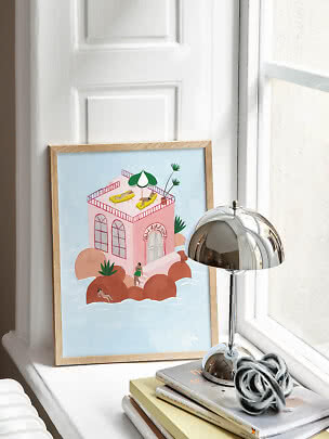 Gerahmte Illustration eines rosa Hauses am Wasser auf einer Fensterbank neben einer Chromlampe