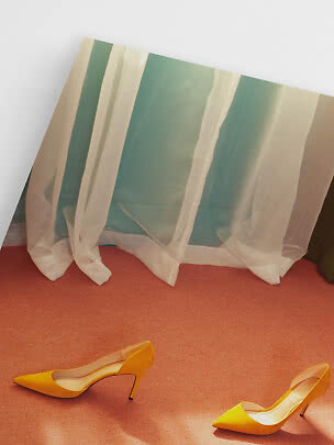 Pair of yellow pointed-toe stiletto pumps on an orange floor by a window with sheer curtains