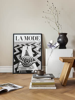 Stylish interior with framed La Mode poster, vase with branches, marble pedestal and stack of books.