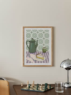 Lámina enmarcada en la pared con cafetera verde, taza y porción de tarta sobre mantel a cuadros
